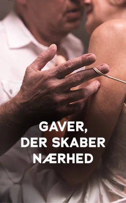 Billedet viser en reklame, hvor en kvinde og en mand står tæt sammen. Manden bærer en hvid skjorte, og kvinden bærer en lys top med tynde stropper. Deres hoveder er tæt på hinanden, og manden trækker med sin hånd kvindens tynde stropper ned ad hendes skulder. Med store, hvide bogstaver står der »Gaver, der skaber nærhed«. Nederst er der en rød knap med den hvide påskrift »Vis mig dem«. Baggrunden er mørk.