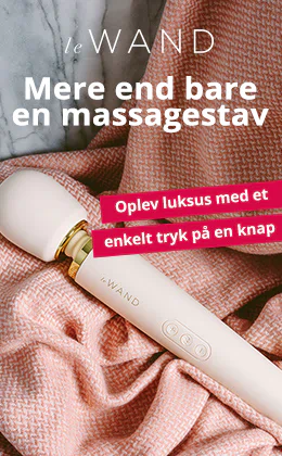 En hvid massagestav af mærket le WAND ligger på et rosafarvet strikket stof foran en grå marmoroverflade. Øverst står der »Mere end en massagestav«, til højre »Oplev luksus med et tryk på en knap«. Nederst er der en rød knap med teksten »Oplev det nu«.